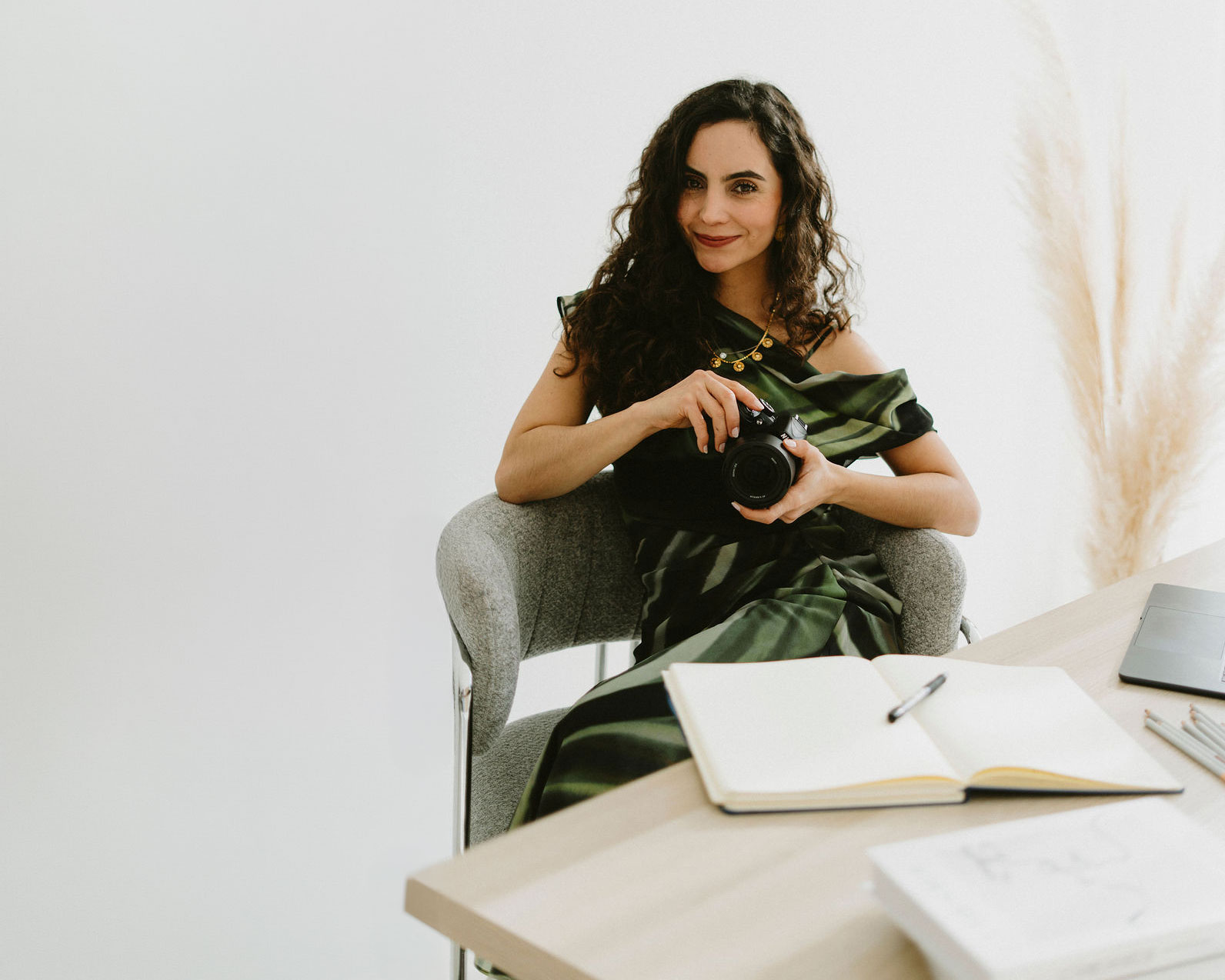 Woman with curly hair sitting in a gray chair holding a camera, with an open notebook and laptop on a desk nearby.