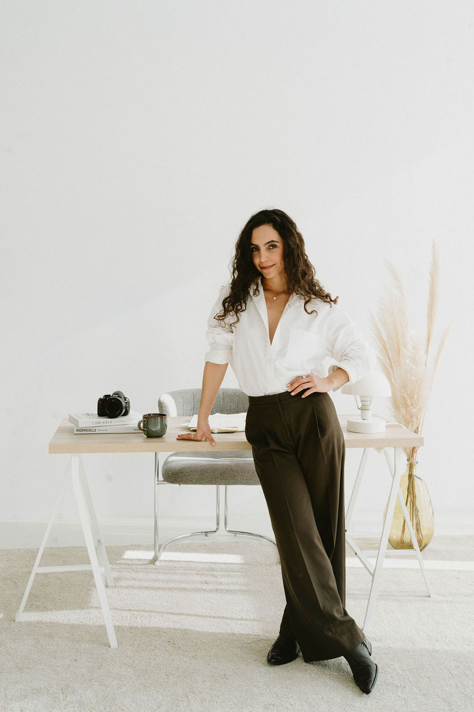 Woman with curly hair leaning on a desk in a bright room, wearing a white shirt and dark trousers.
