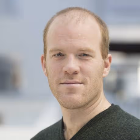 Close-up portrait of a man with short light brown hair and a slight smile, wearing a dark green sweater.