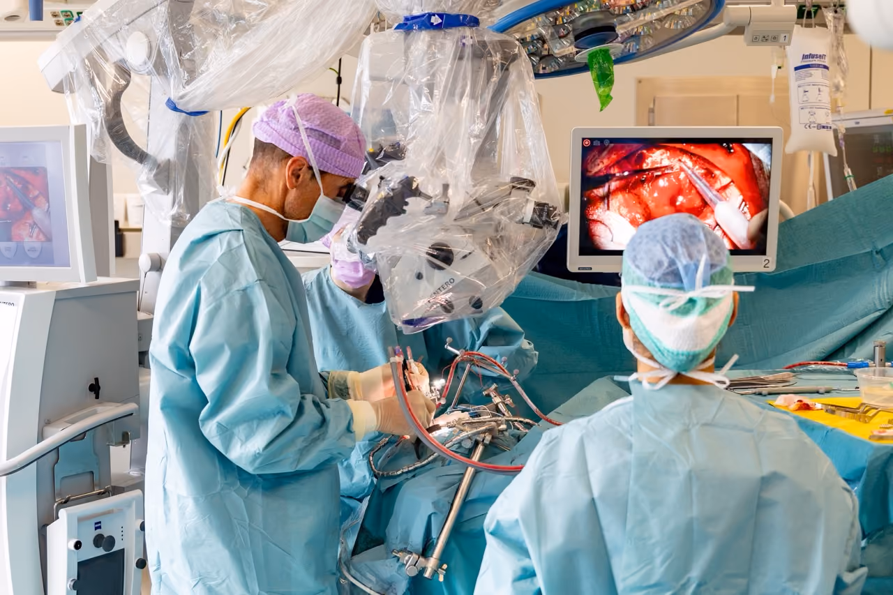 Surgeons in an operating room use a surgical microscope and watch a monitor showing the procedure.