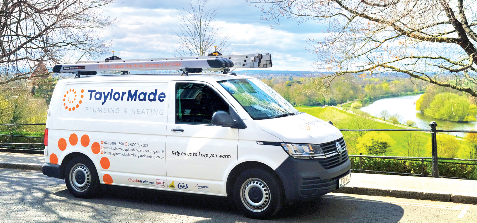 White TaylorMade Plumbing & Heating van parked by roadside with river and green trees in background.