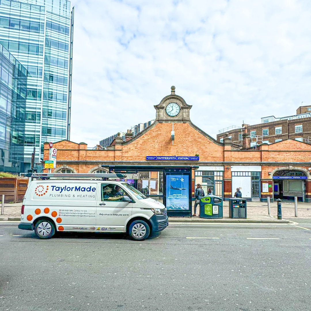 TaylorMade plumbing van parked in front of Hammersmith Station building with clock and people nearby.