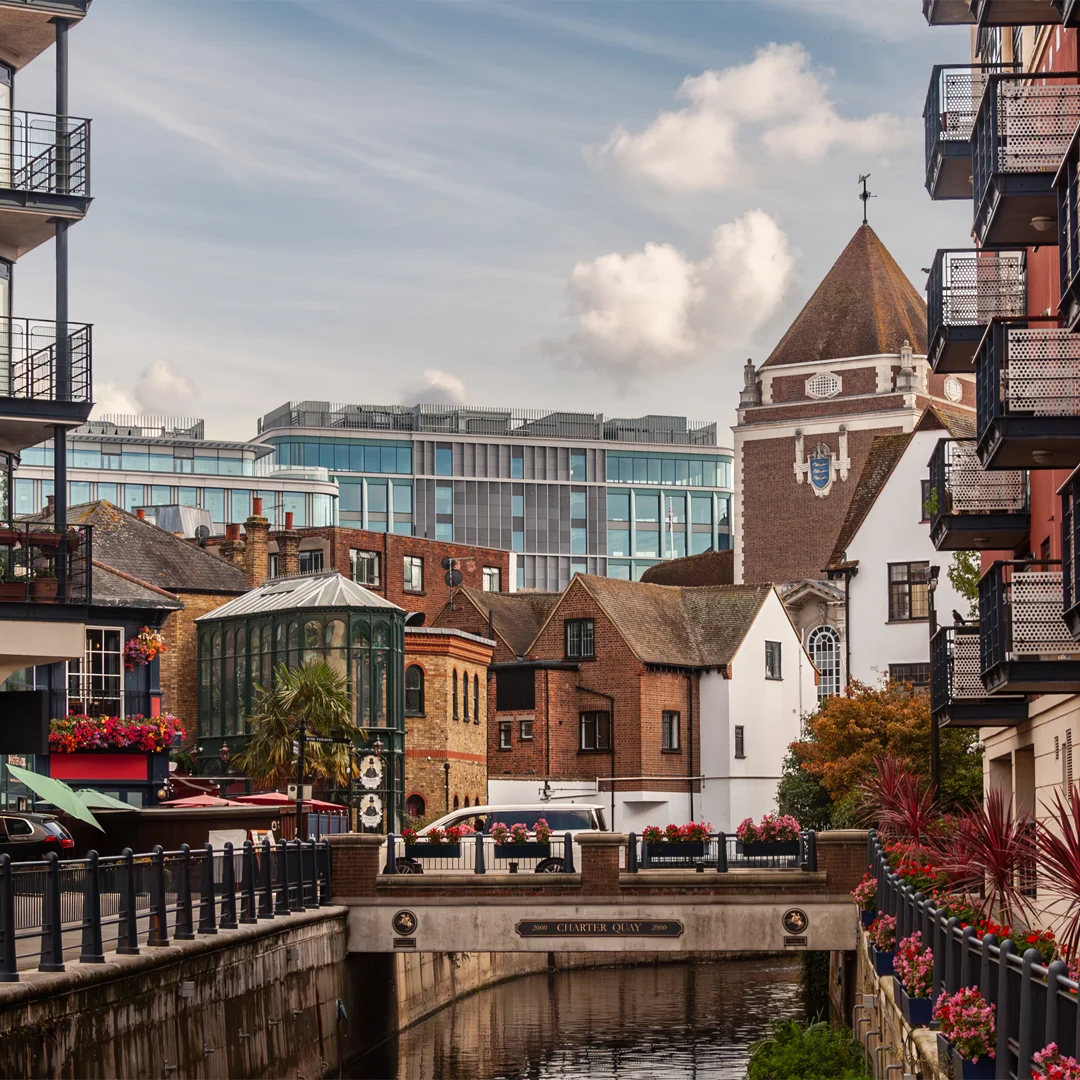 River canal with stone bridge, historic and modern buildings, flowers, and blue sky with clouds.