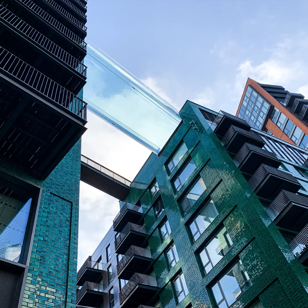 View looking up at modern green and black buildings connected by a glass skybridge under a blue sky.