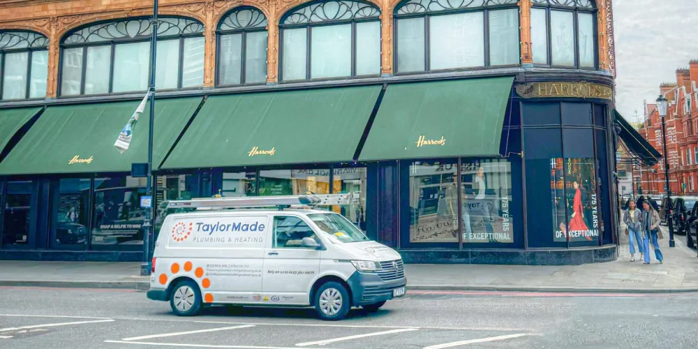 White TaylorMade Plumbing & Heating van parked beside road with scenic river and trees in background.