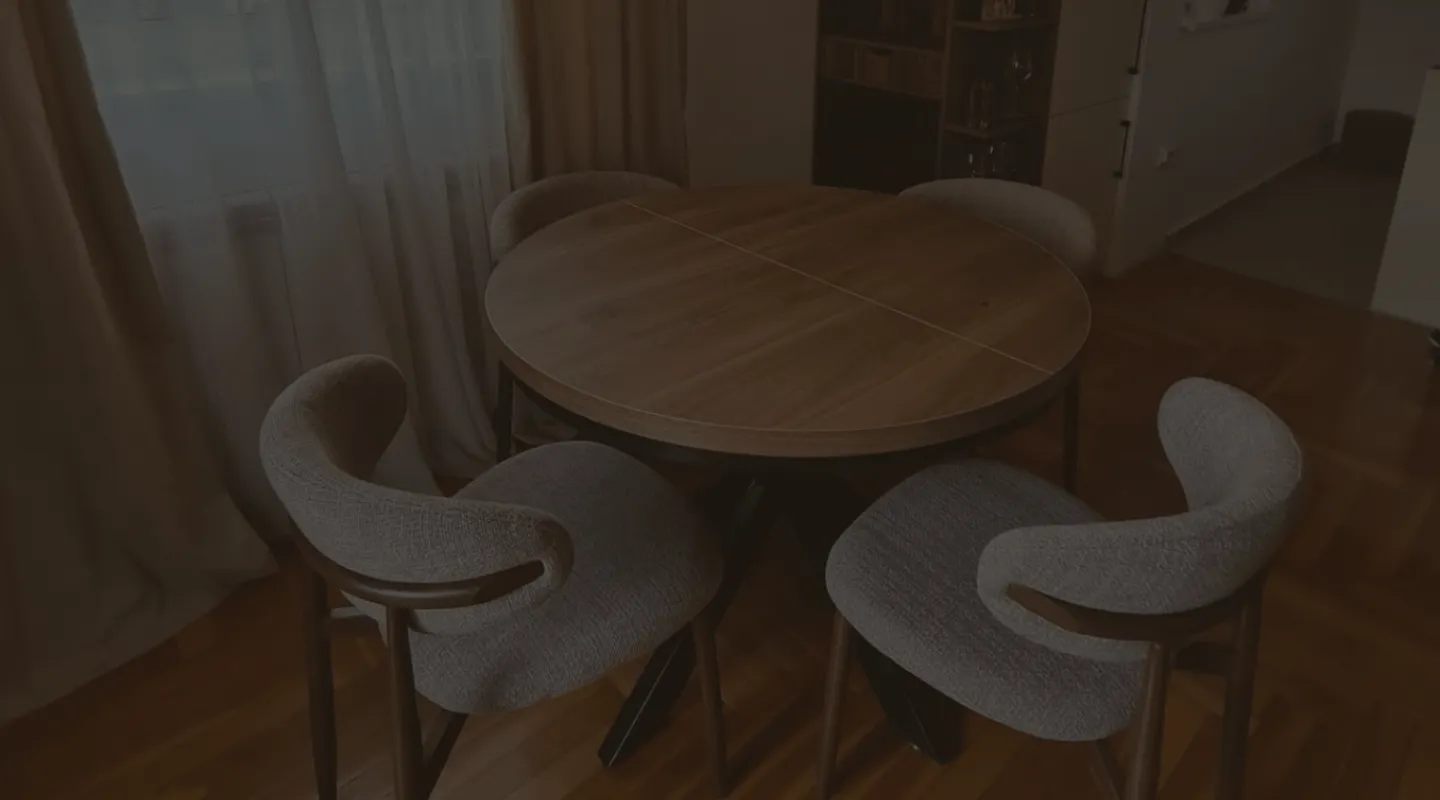 Okrugli drveni trpezarijski sto sa četiri zakrivljene, udobno tapacirane stolice, smešten u enterijer sa toplim drvenim podom i prozorom sa zavesama.