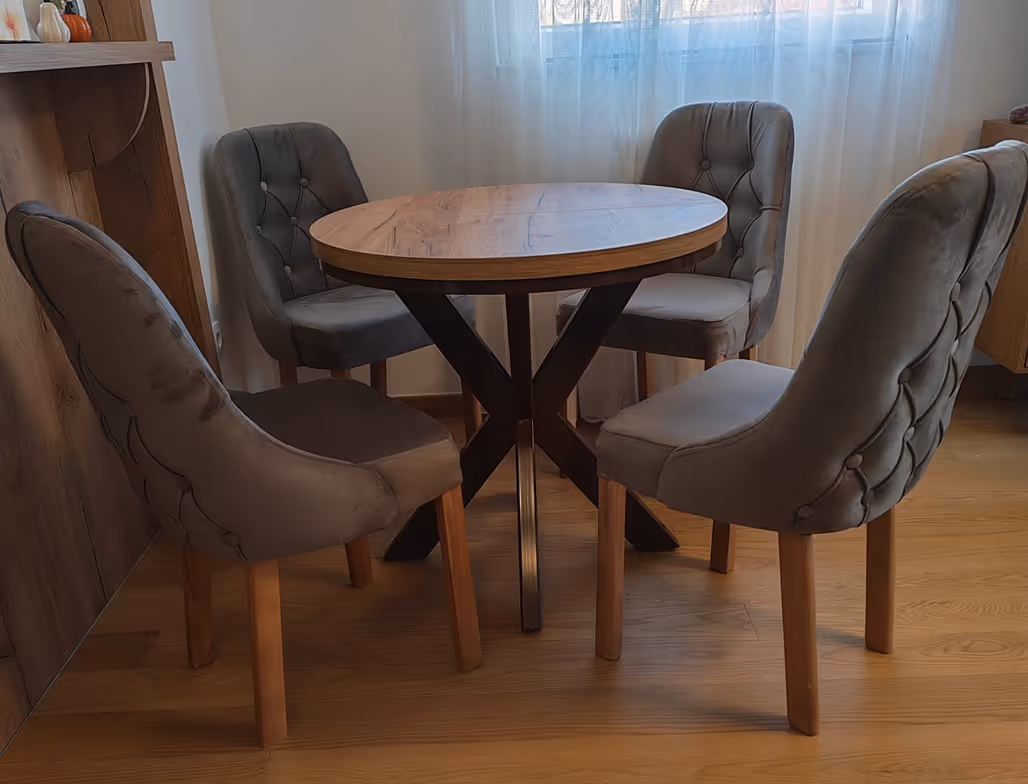 Okrugli drveni sto sa crnim metalnim postoljem, okružen sa četiri tapacirane stolice u senf žutoj nijansi, na toplom drvenom podu.