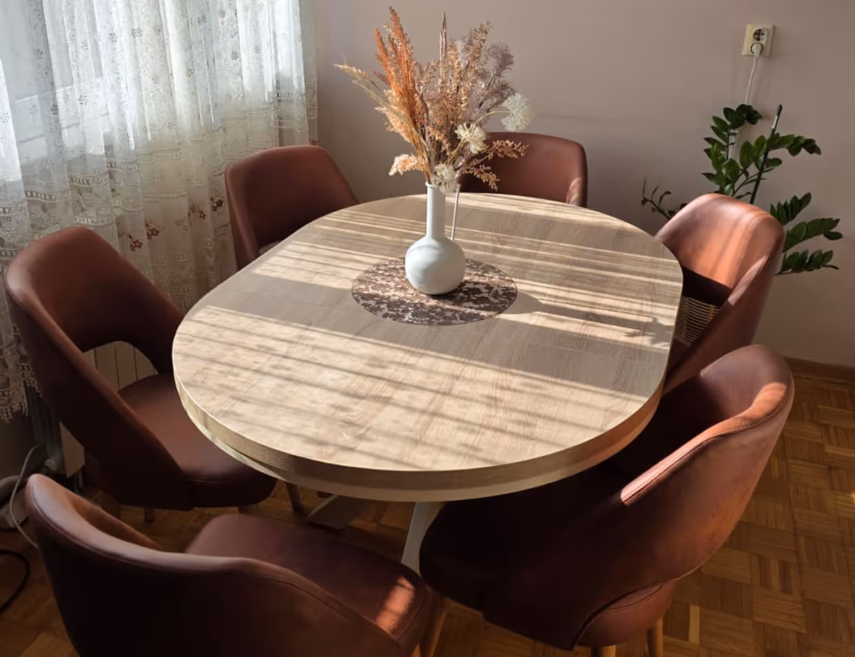 Okrugli drveni sto sa crnim metalnim postoljem, okružen sa četiri tapacirane stolice u senf žutoj nijansi, na toplom drvenom podu.