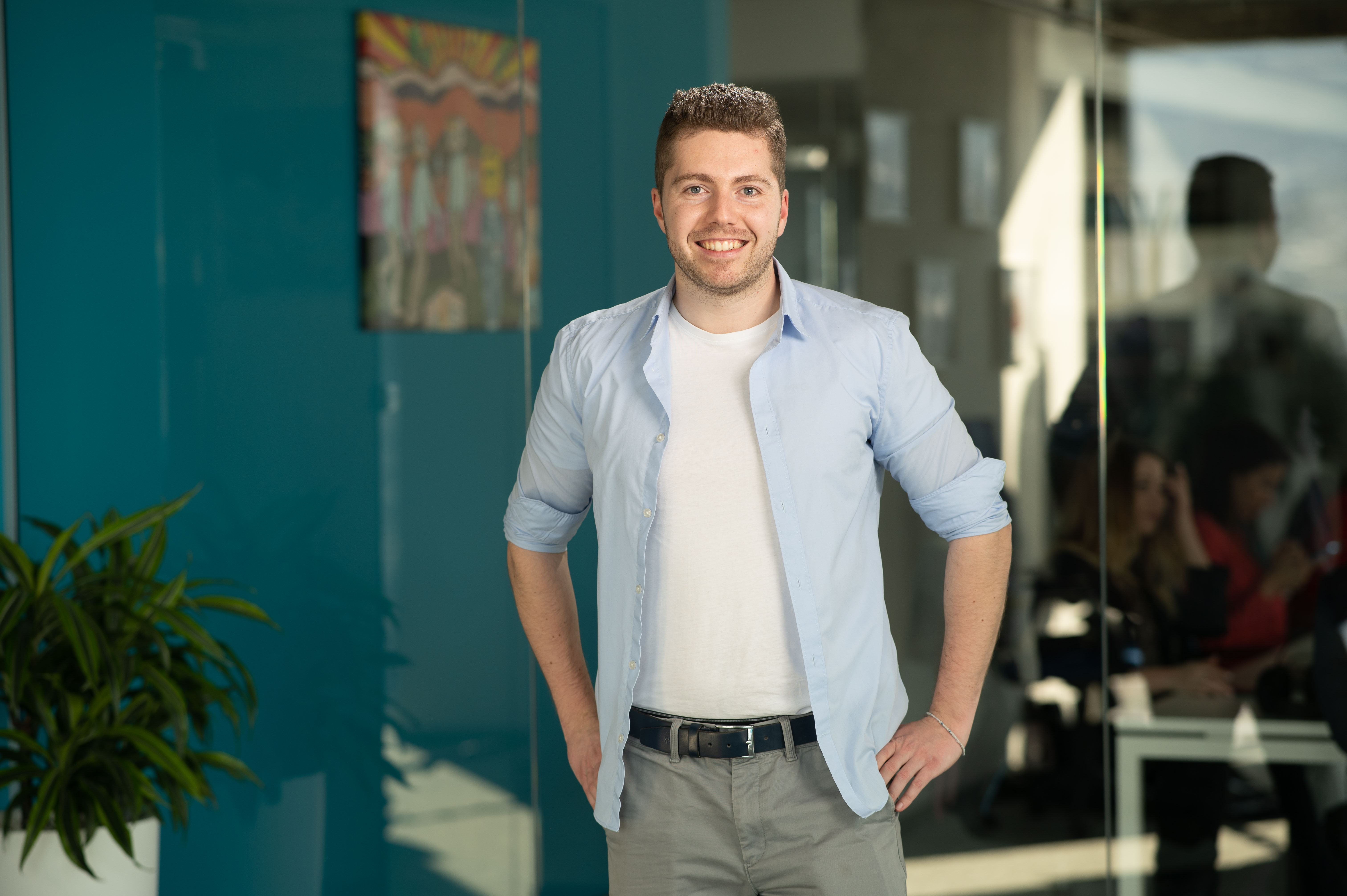 Smiling man with short brown hair wearing a light blue shirt over a white t-shirt, standing with hands on hips in an office with glass walls and plants.