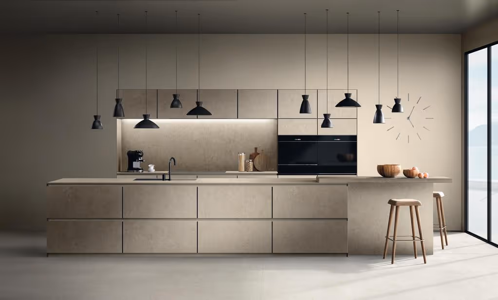 Moderne minimalistische keuken met betonnen werkblad, zwarte hanglampen, ingebouwde oven, koffiezetapparaat en twee houten krukken naast een raam.