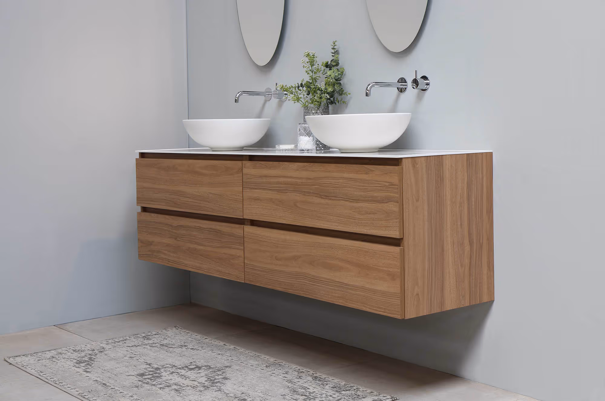 Moderne badkamermeubel met zwevende houten kast, twee witte waskommen, ronde spiegels en een klein groen plantenarrangement.