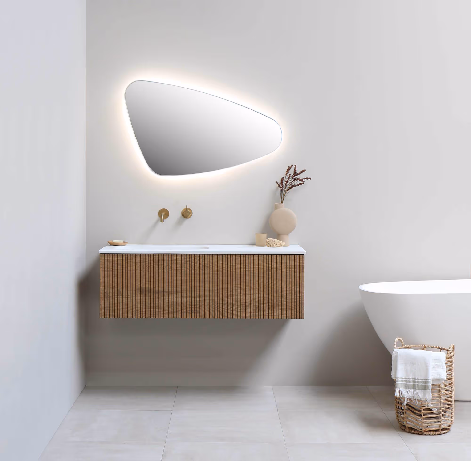Minimalistisch badkamerinterieur met hangende houten wastafel, asymmetrische verlichte spiegel en vrijstaand wit bad met mand voor handdoeken.