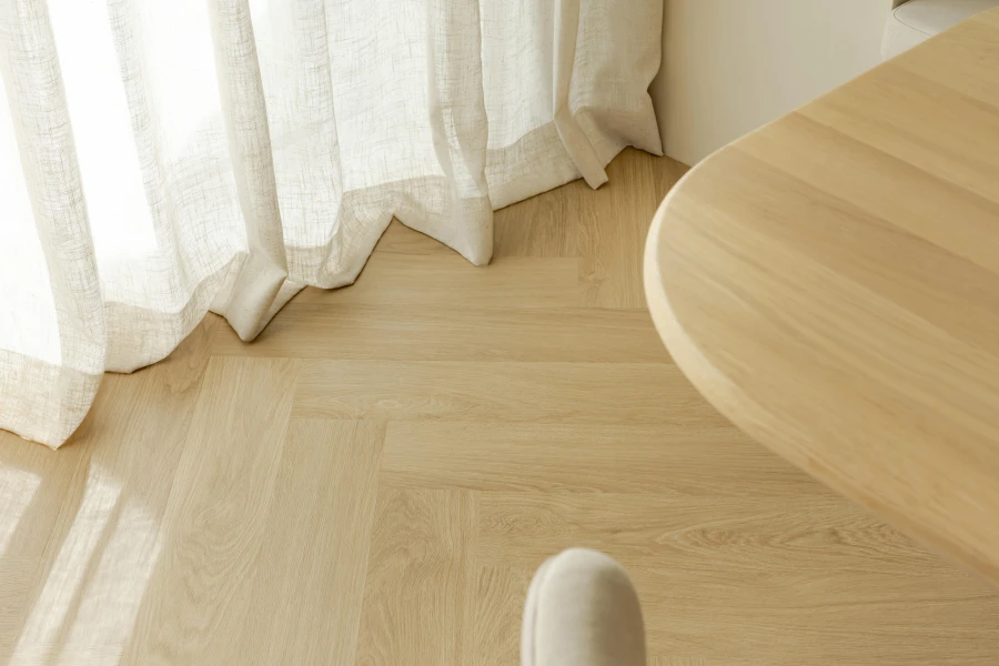 Licht houten visgraatvloer met zonlicht door witte gordijnen en een afgeronde houten tafel.