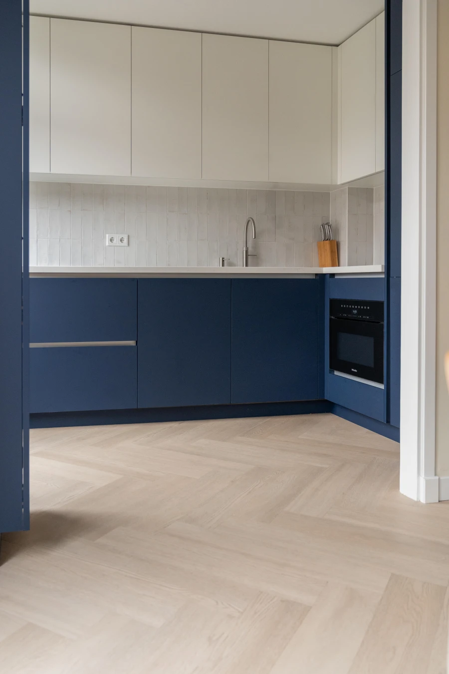 Moderne keuken met donkerblauwe onderkasten, witte bovenkasten, een lichte houten visgraatvloer en een inbouwoven.