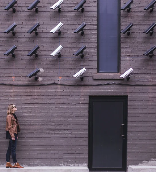 Une femme lève les yeux vers un mur en briques couvert de caméras de surveillance, illustrant les enjeux du RGPD et de la cybersécurité.
