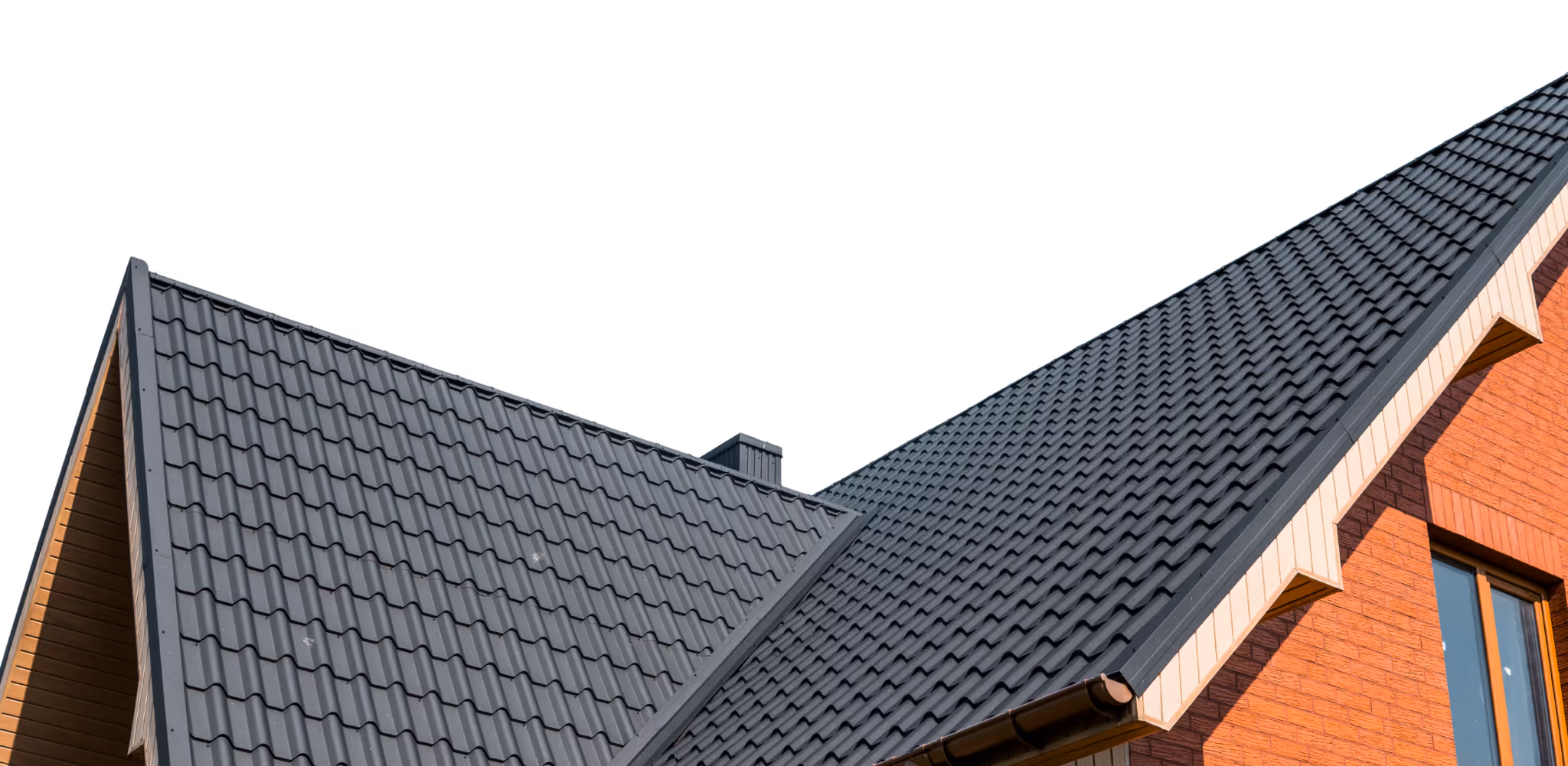 Close-up view of a house roof with dark gray metal tiles and a red brick wall with a window.