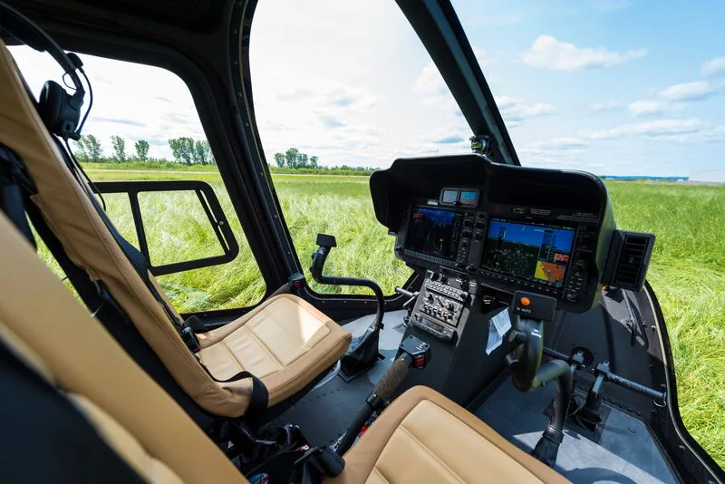Helicopter cockpit with control panels and two beige seats, set against a green field backdrop.