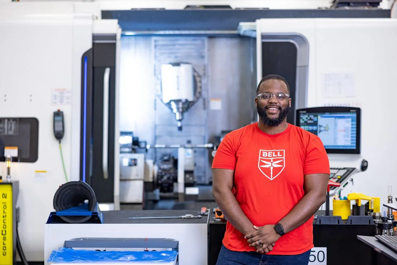 A man in a red Bell T-shirt stands in a manufacturing facility with machinery in the background.