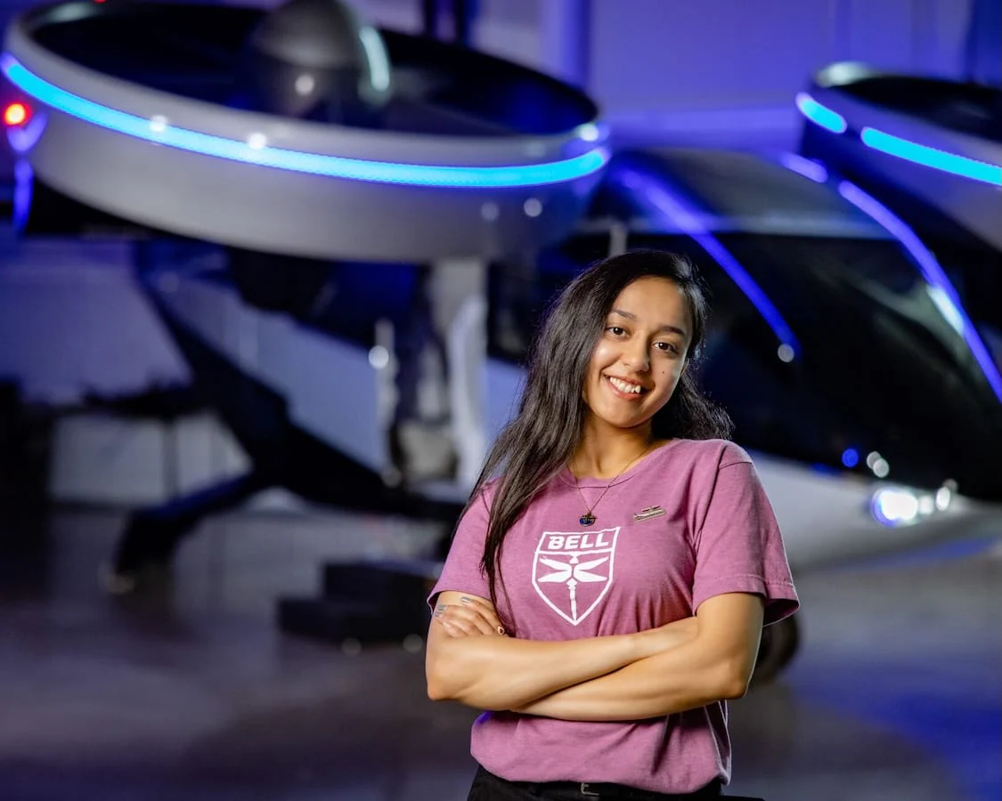A woman in a purple shirt stands confidently in front of a futuristic aircraft design.