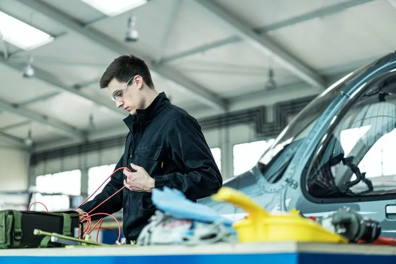 Young man in safety glasses working on electrical equipment in an aircraft repair workshop.