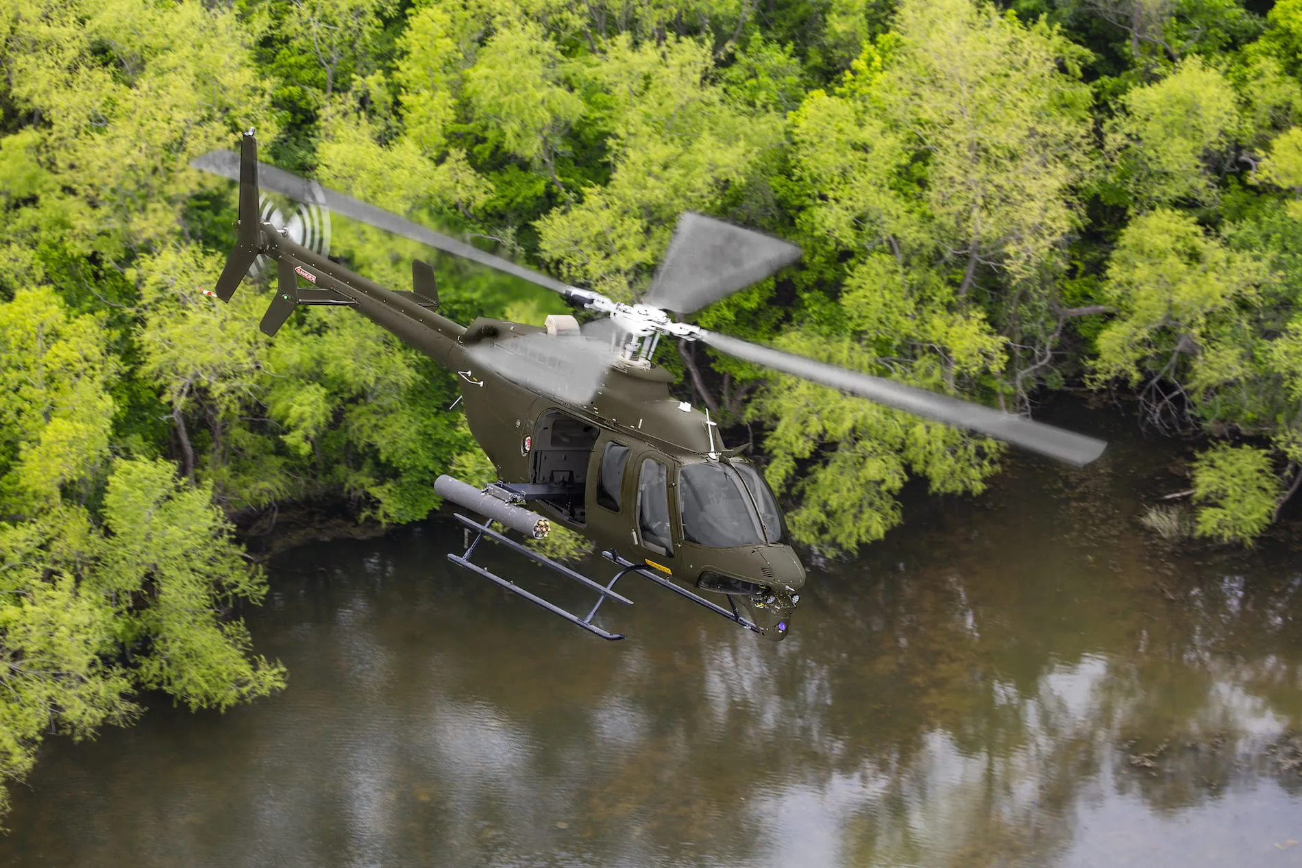 Military helicopter flying over a forested area with a calm water surface below.