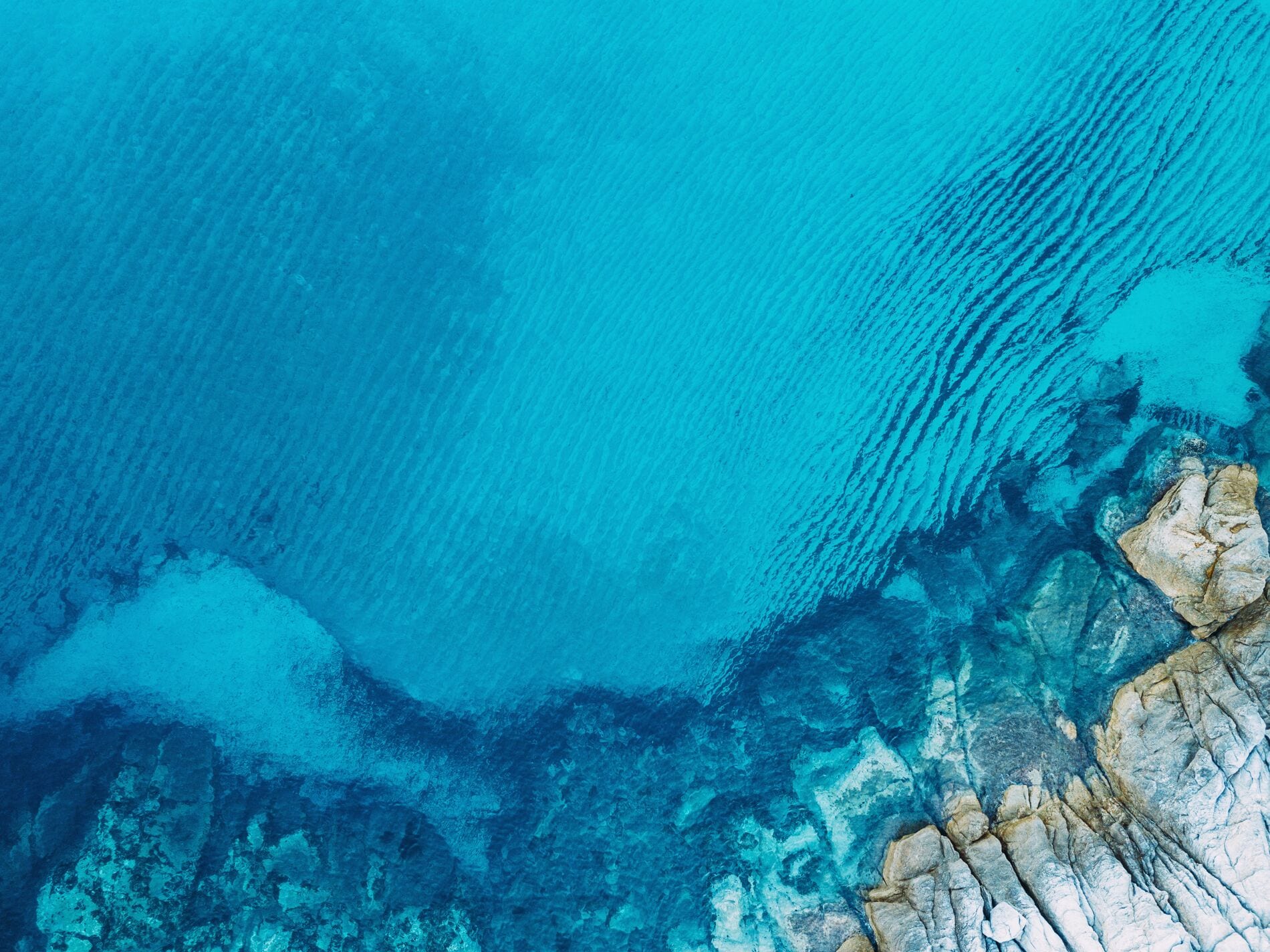 Aerial view of clear turquoise water with rocky shoreline and ripples visible in the water.