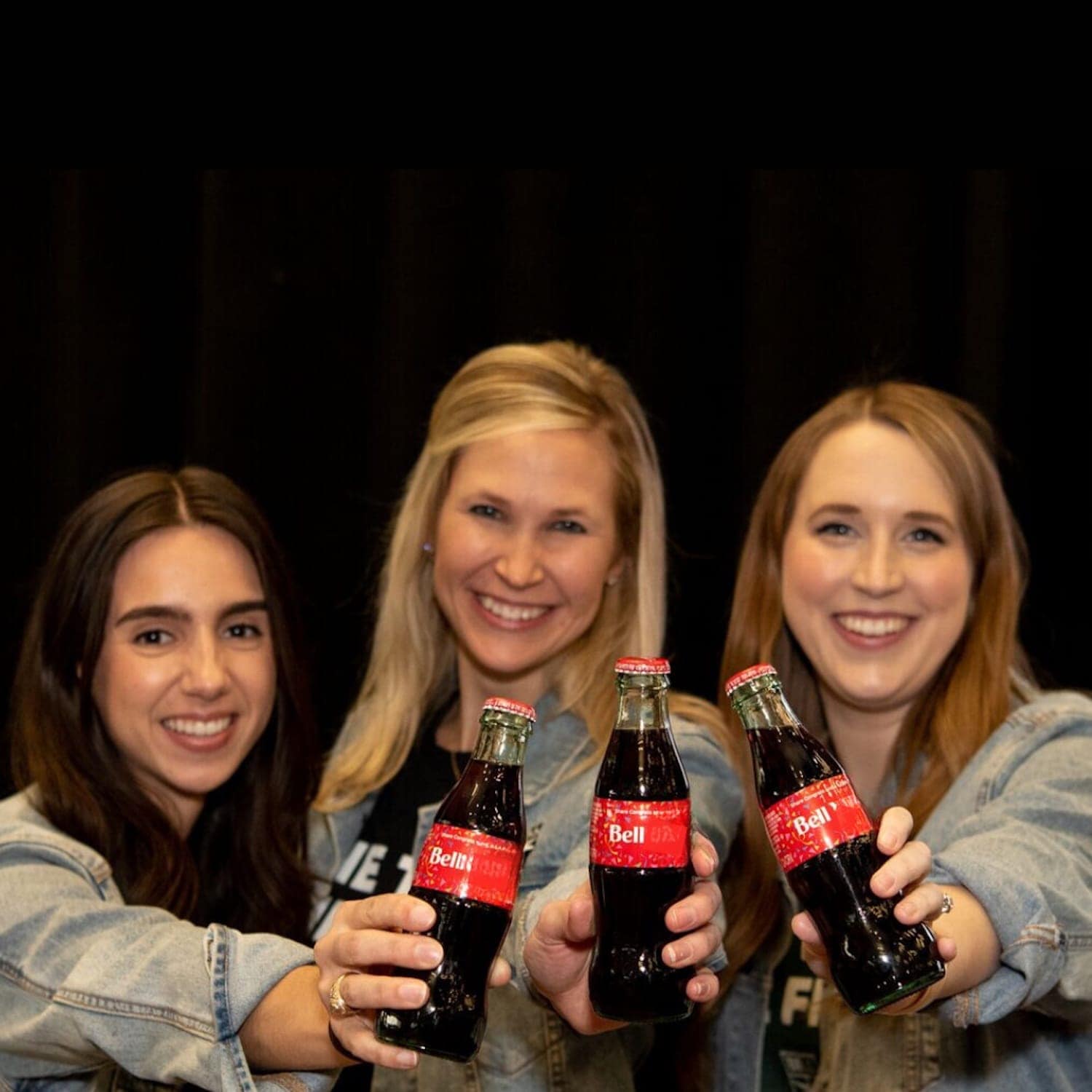 Three women in denim jackets hold up Coca-Cola bottles, smiling brightly at the camera.