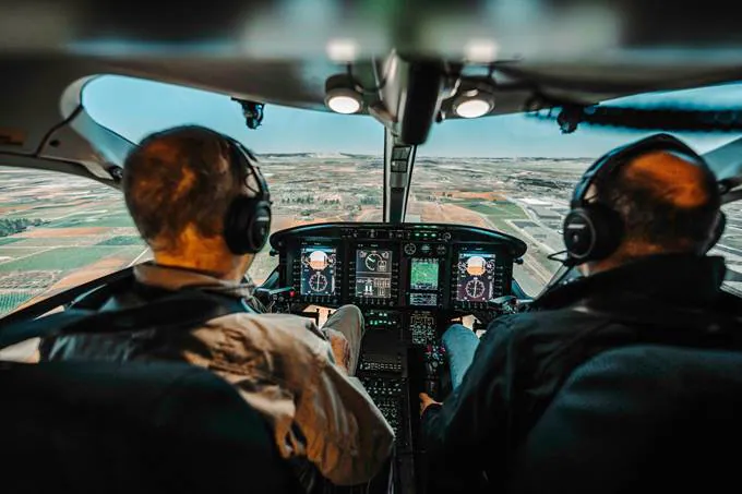 Two pilots in a helicopter cockpit with clear views of the landscape and control panels.