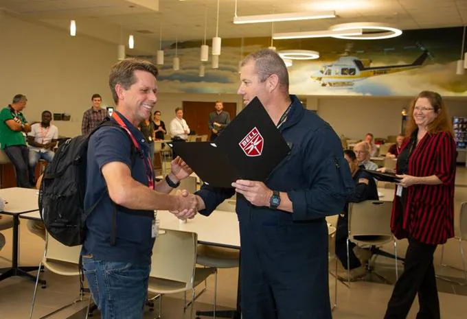 Two men shake hands at a table in a modern room; one holds a folder, people are seated in
