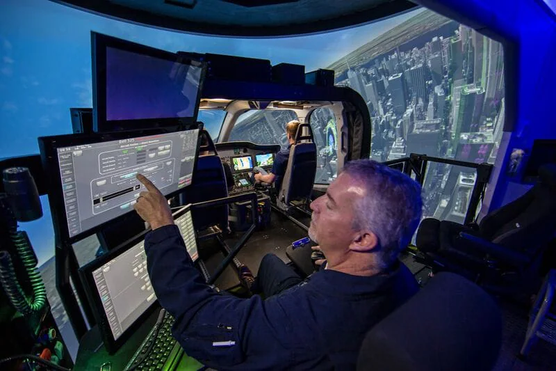 A pilot operates a flight simulator with multiple screens and a cityscape backdrop.