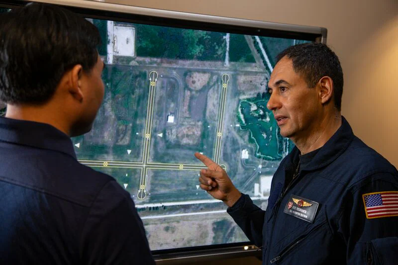 Two men in military uniforms review an aerial map of an airstrip, discussing details and plans.