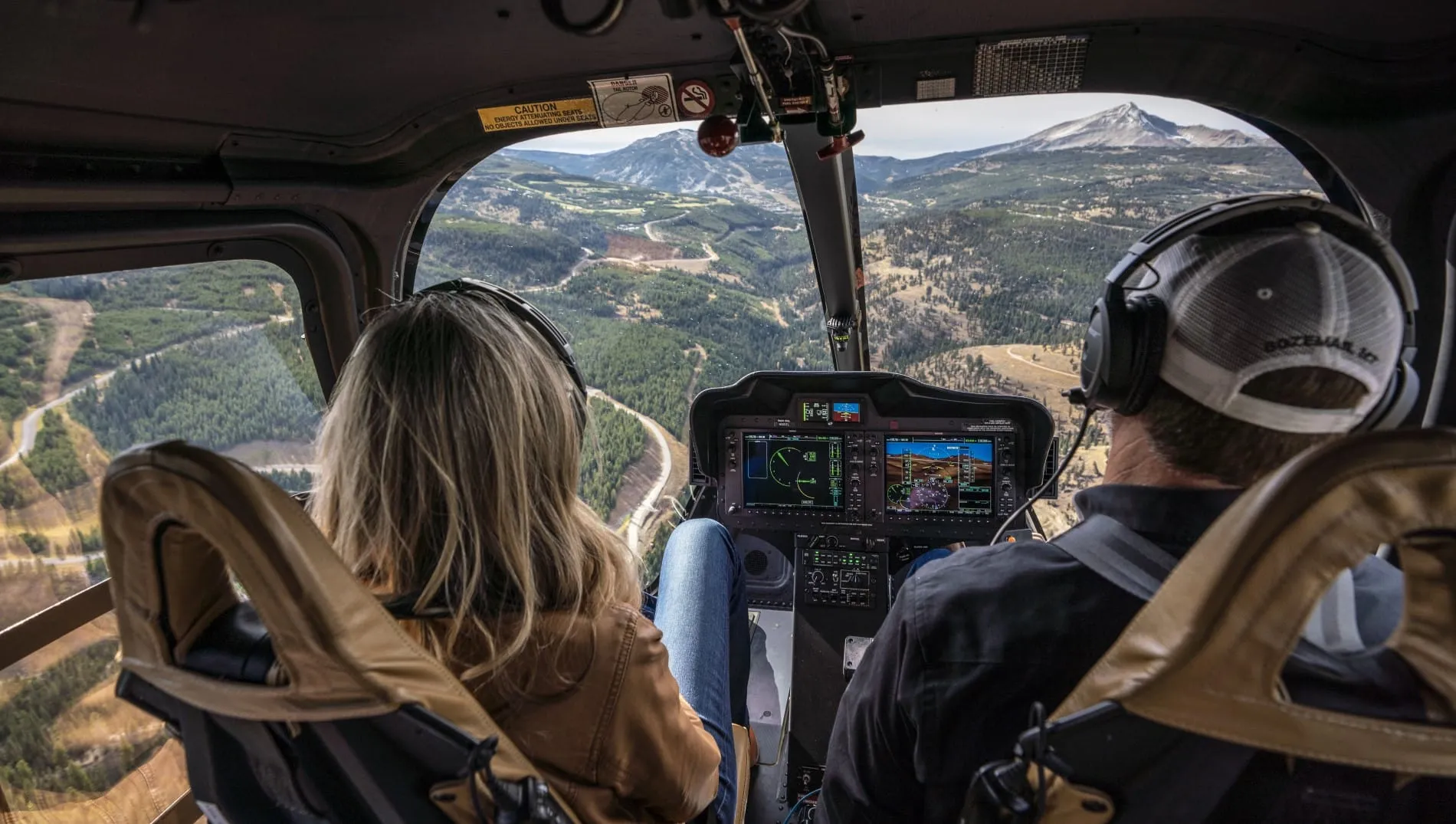 Helicopter cockpit view with two pilots, mountains and forests visible through the windshield.