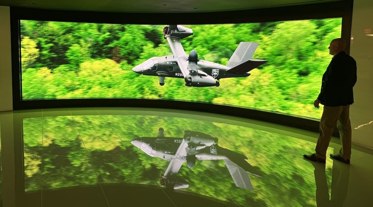 A man walks by a large display showing a military drone flying over green foliage.
