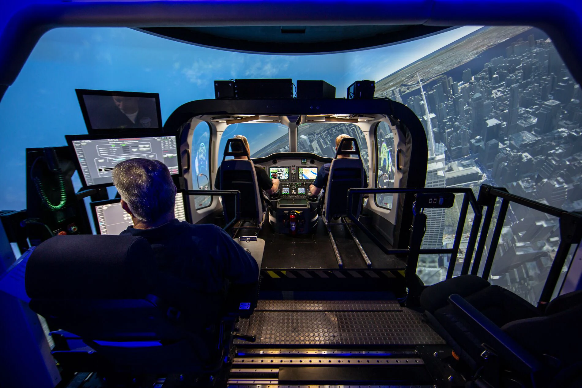 A pilot sits in a flight simulator with panoramic digital cityscape displays.
