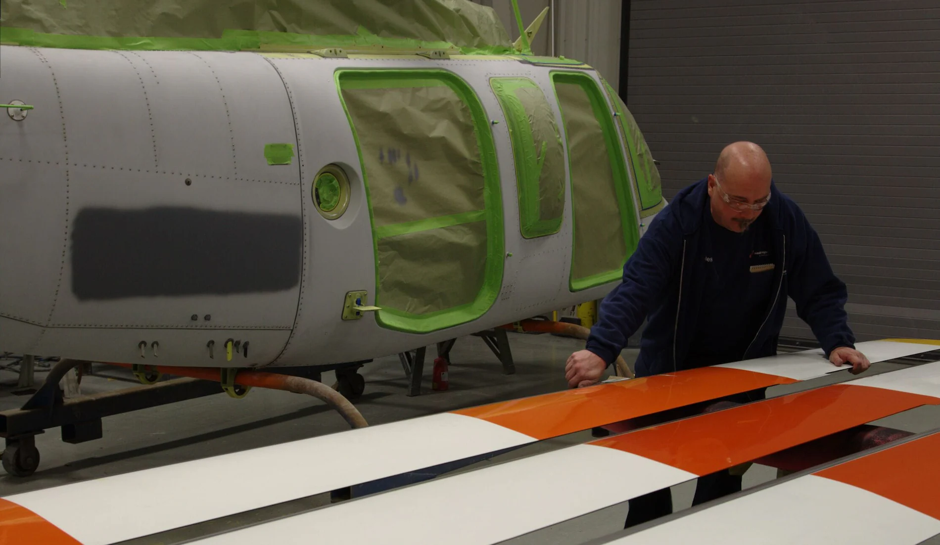 A person works on a helicopter fuselage with green accents and painted rotor blades in a workshop.