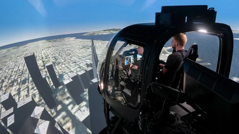 A person piloting a helicopter simulator with a panoramic cityscape projection on screens.
