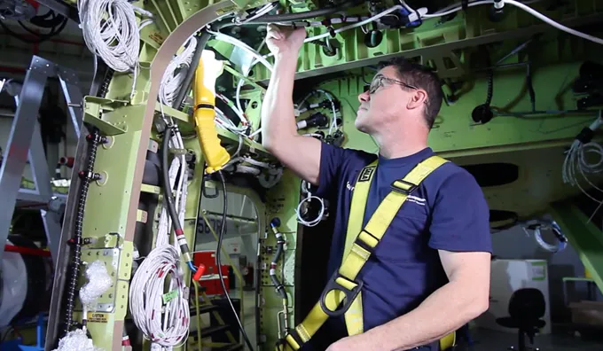 A technician in safety gear repairs wiring inside an aircraft fuselage. Tools and equipment are