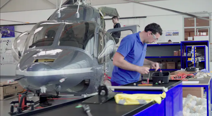 Mechanic working on a helicopter in a hangar with tools and equipment around.