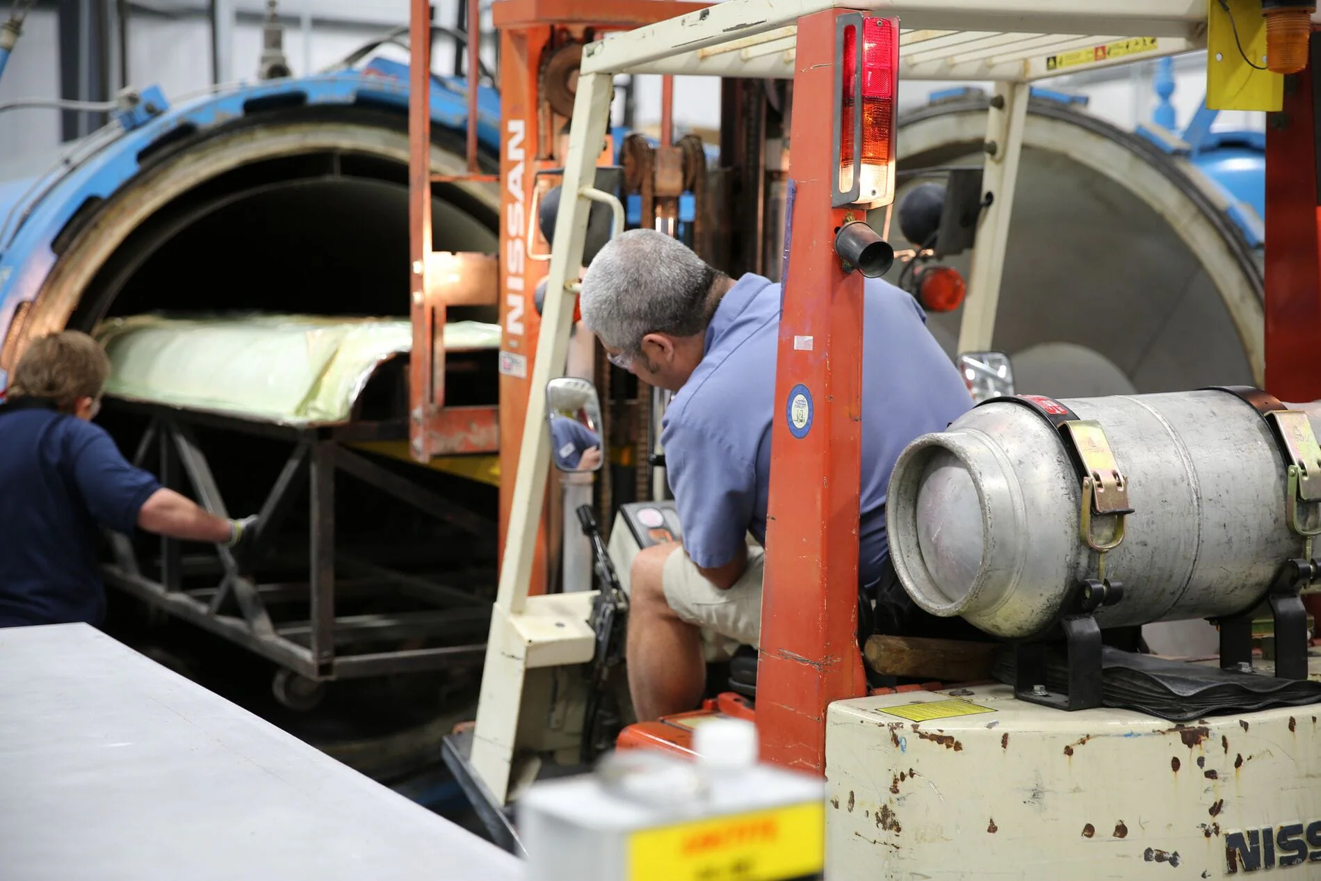 Two workers in a warehouse operate machinery near large cylindrical equipment.