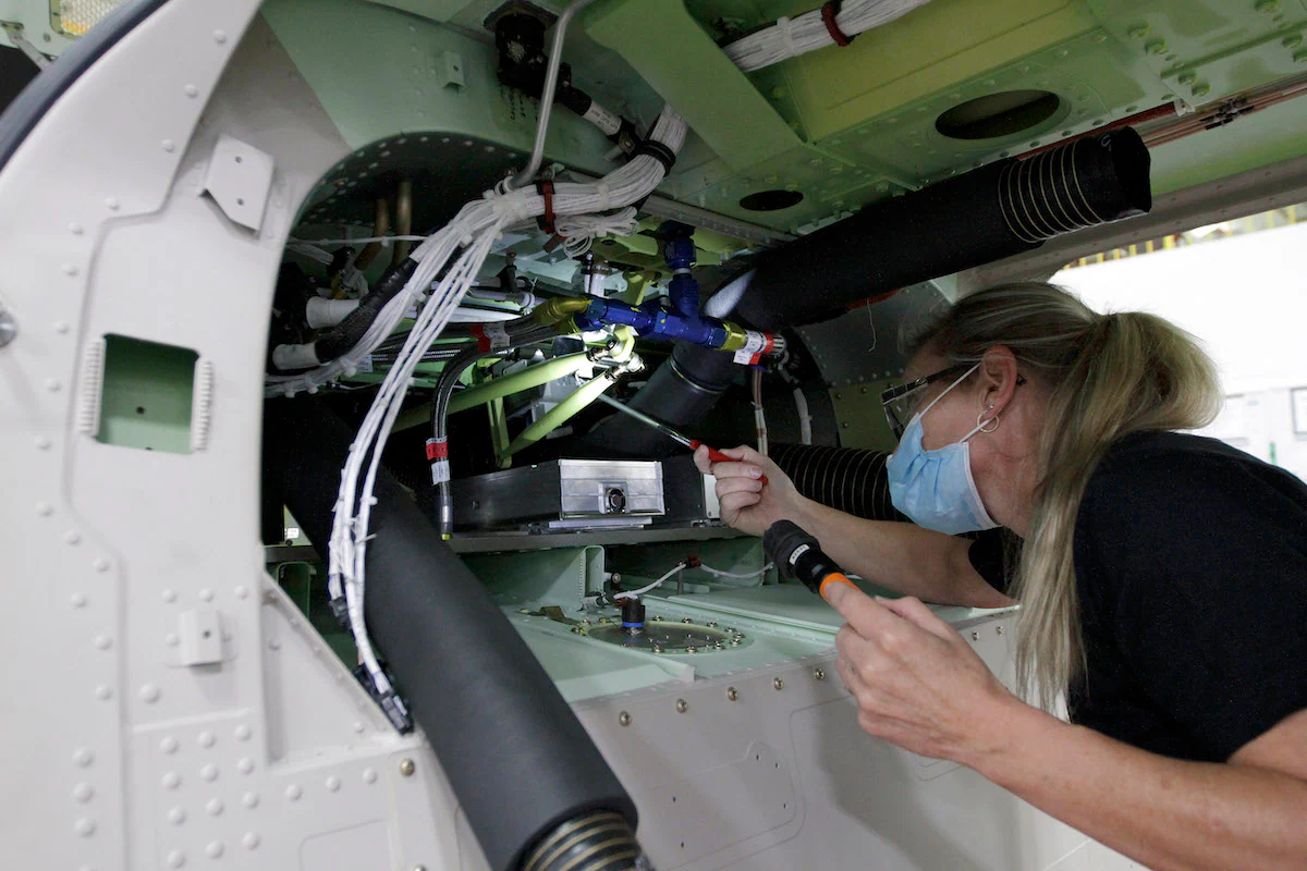 A technician inspects aircraft components inside a fuselage, using a flashlight and tools.