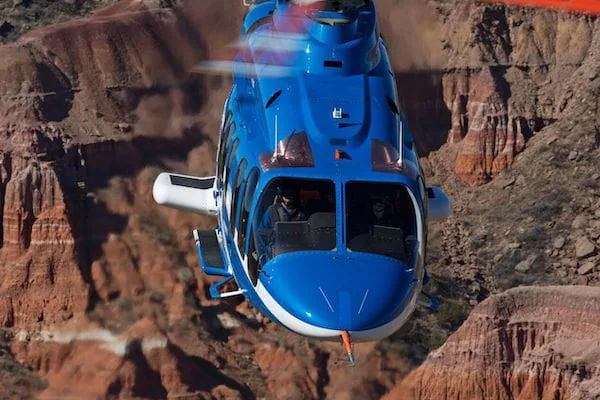 A blue helicopter flies over rocky, rugged terrain with steep cliffs and canyons.