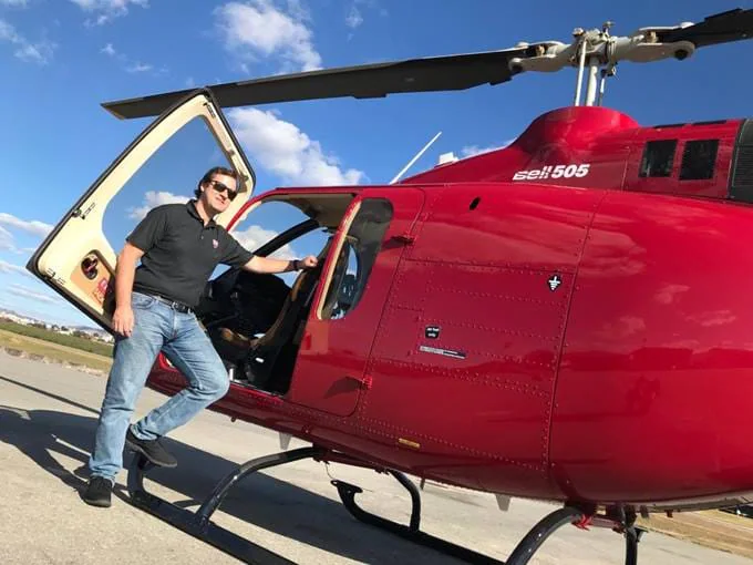 A man stands next to a red helicopter with the door open, smiling against a blue sky.