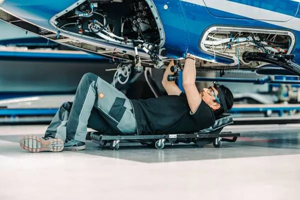 A technician is lying on a dolly, working on the underside of an aircraft. Tools and wires are