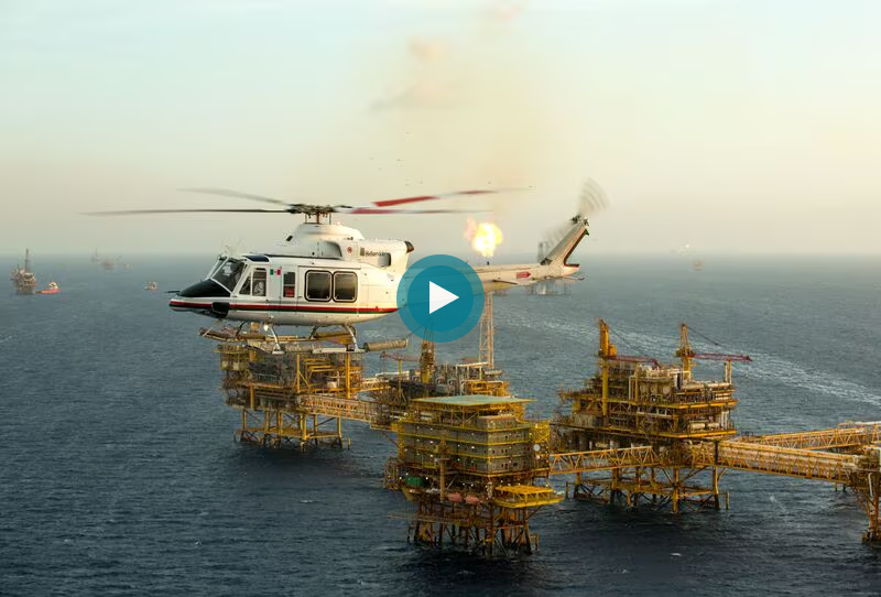 Helicopter flying near an offshore oil platform with explosions in the background at sunset.