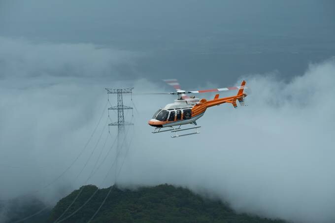 A helicopter flies above a mountainous area, surrounded by clouds and near power lines.