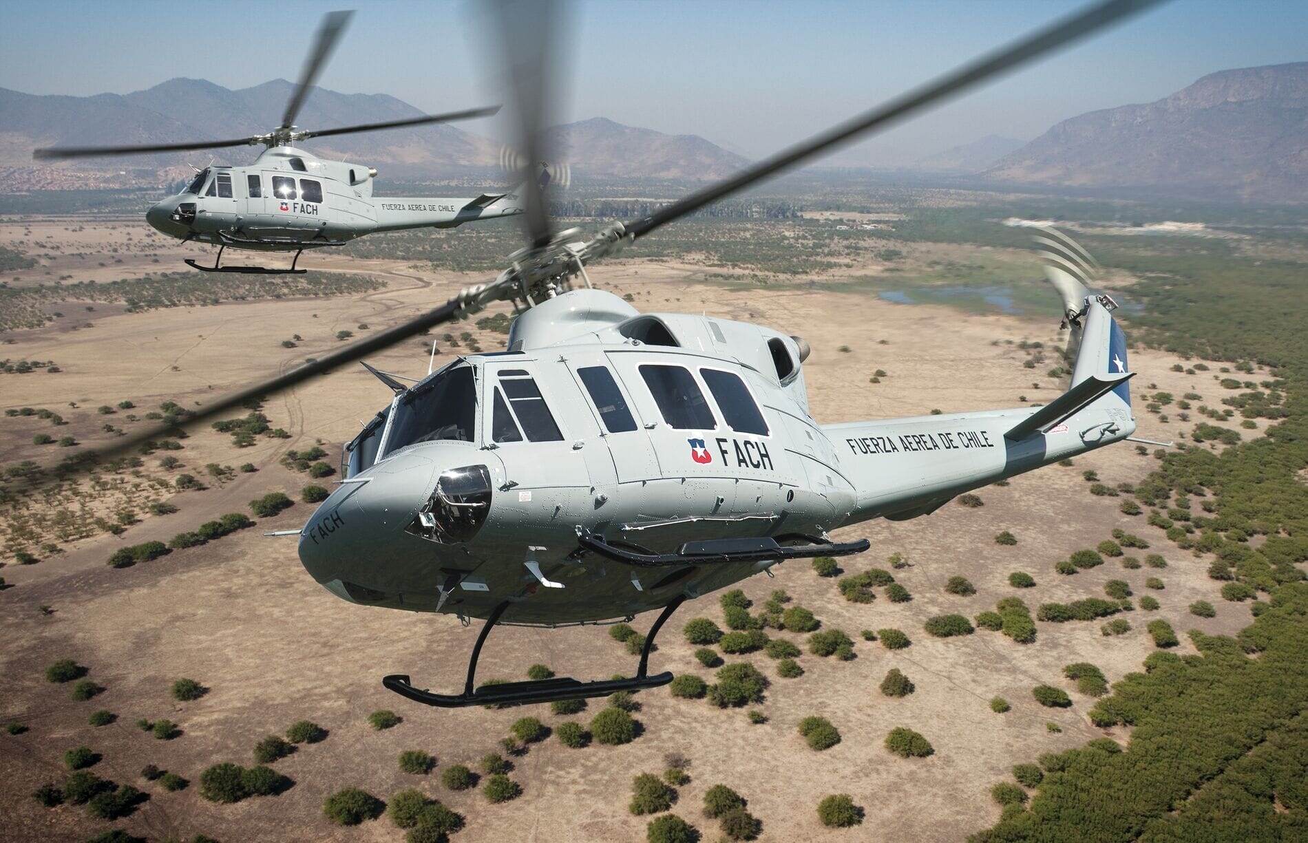 Two military helicopters fly over a dry, mountainous landscape, surrounded by sparse vegetation.