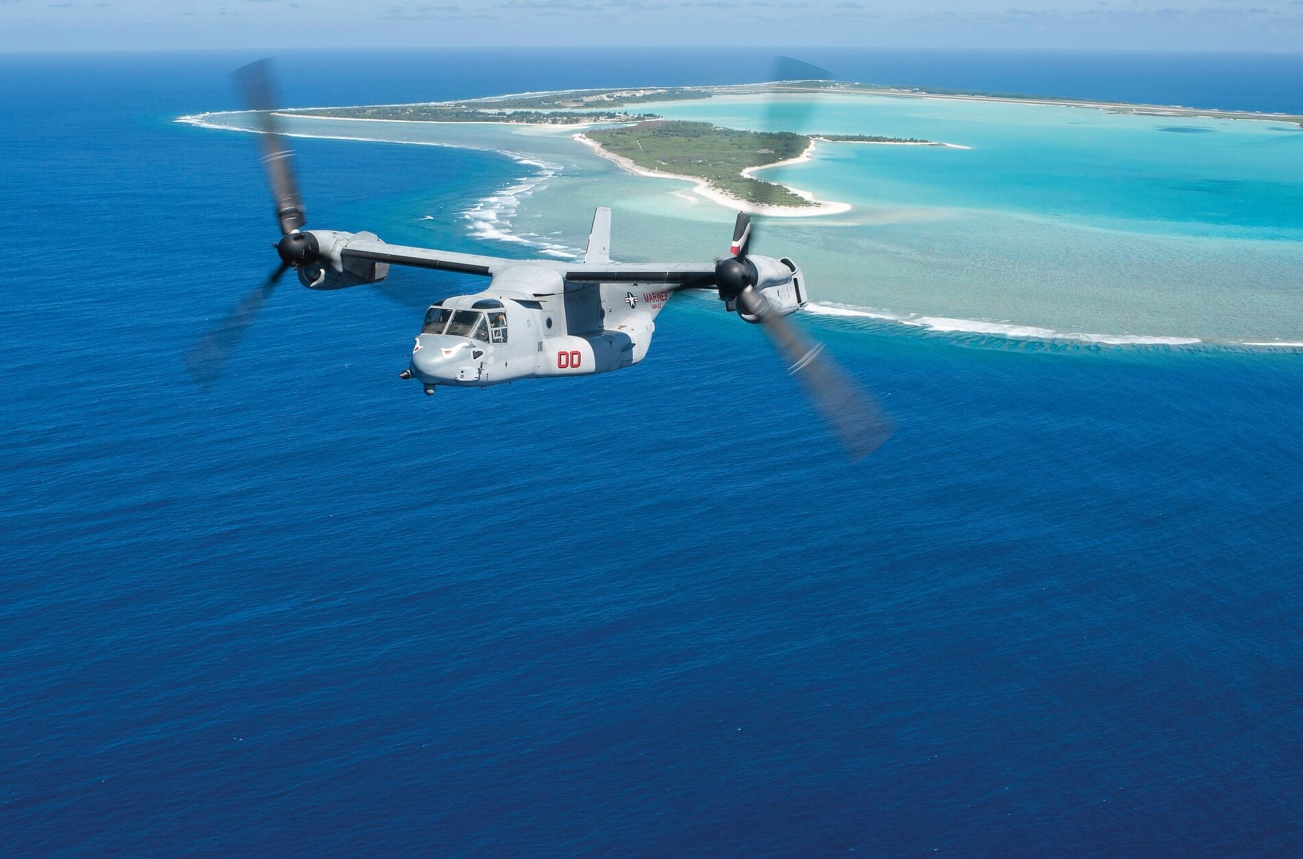 Military aircraft flying over a tropical island with turquoise waters and sandy beaches.