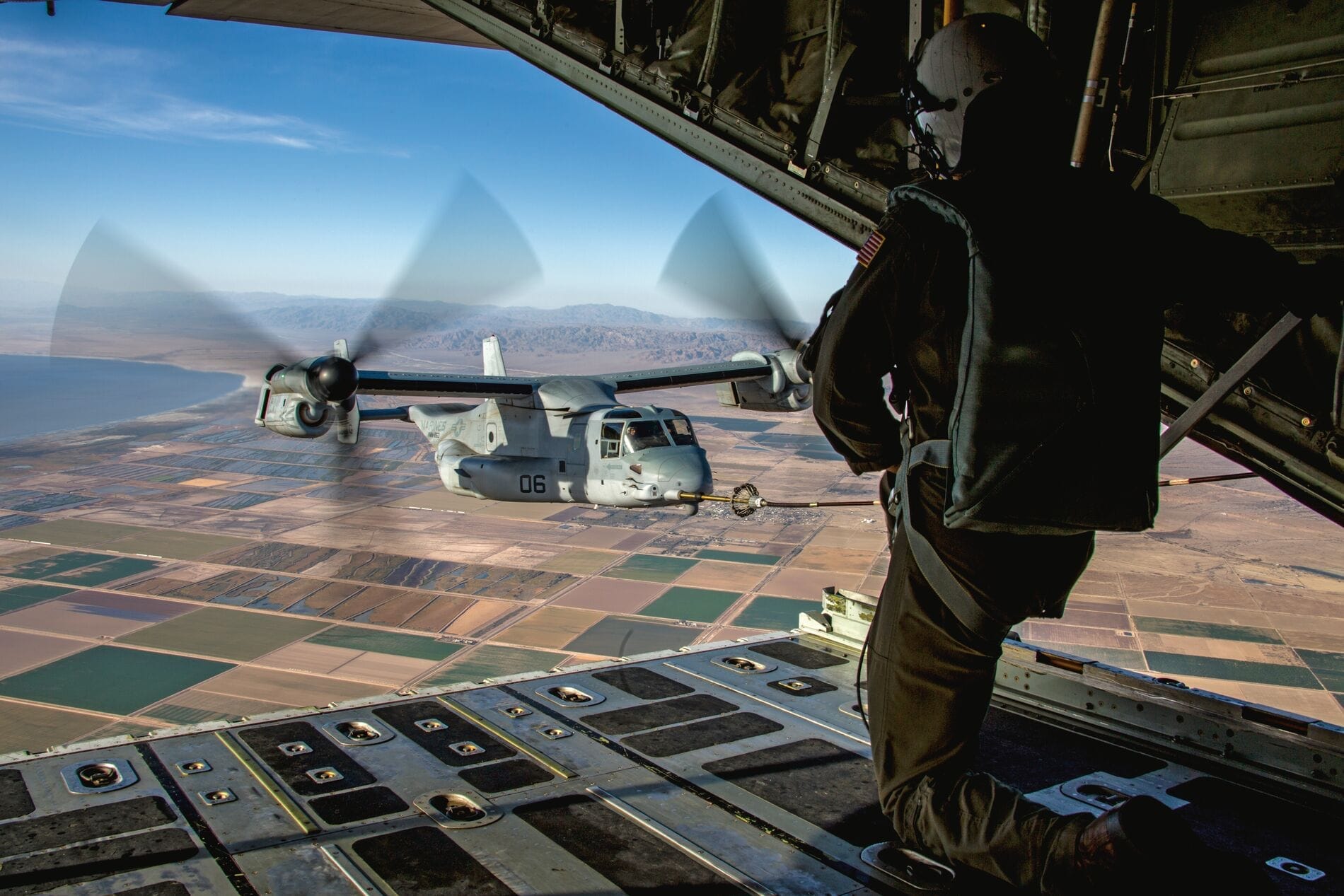 Military personnel surveys landscape from an open cargo door of a transport aircraft; another aircraft flies by.