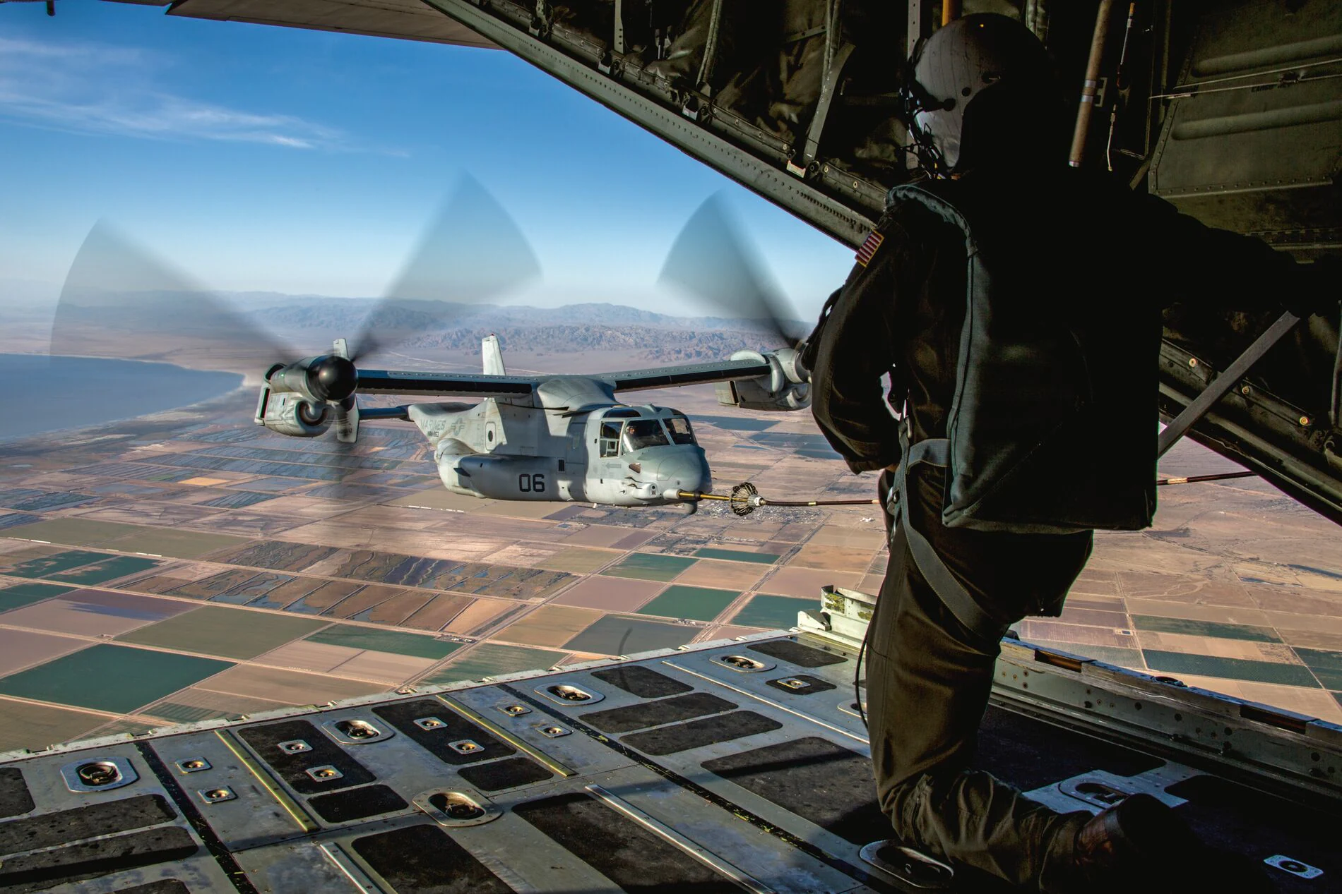 A military aircraft is viewed in flight from the open ramp of another aircraft. Fields are below.