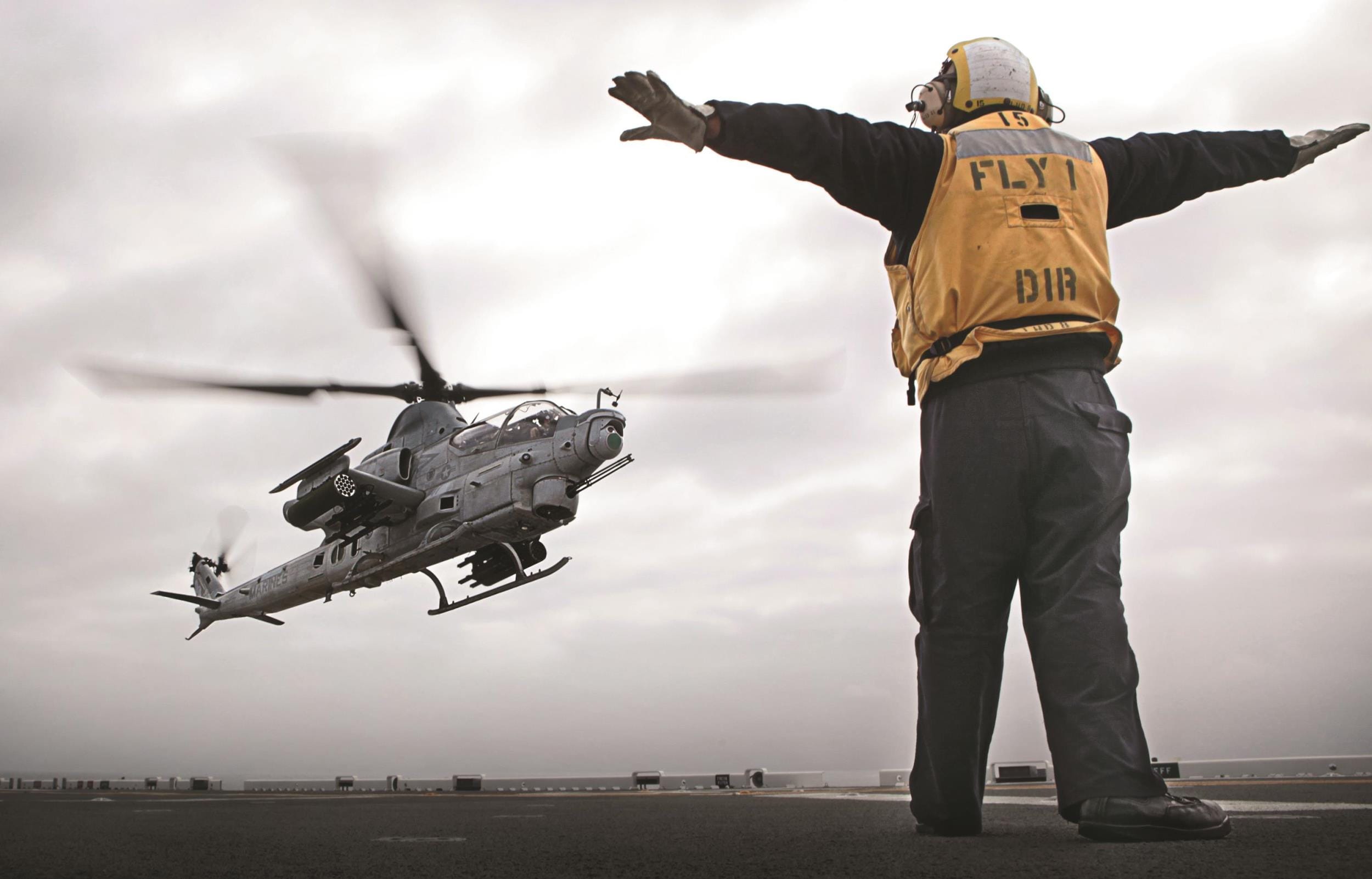 A military helicopter takes off from a naval ship while a crew member signals with arms raised.